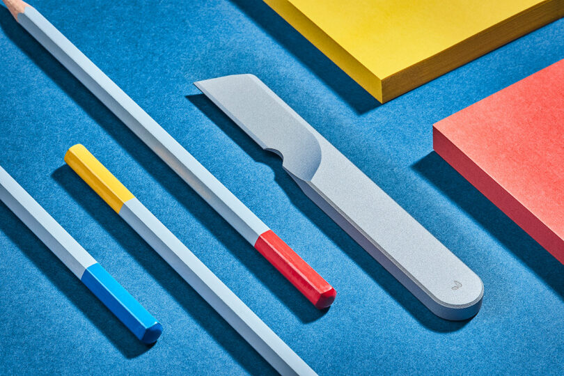 Various desk supplies including two colored pencils, a Task Knife from Grovemade, and yellow and red sticky notes on a blue surface.