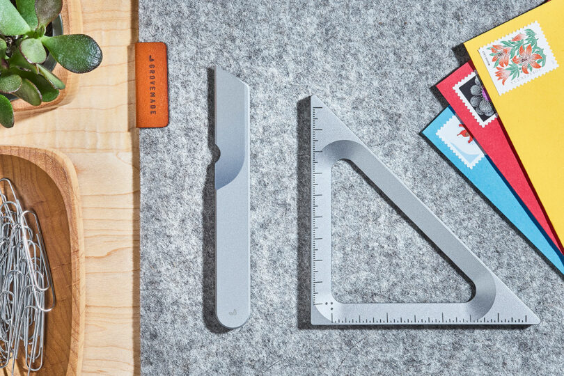 A desk with a utility Task Knife from Grovemade, a metal triangle ruler, a small wooden tray with paper clips, a plant, and a few colorful envelopes with stamps on a grey surface.