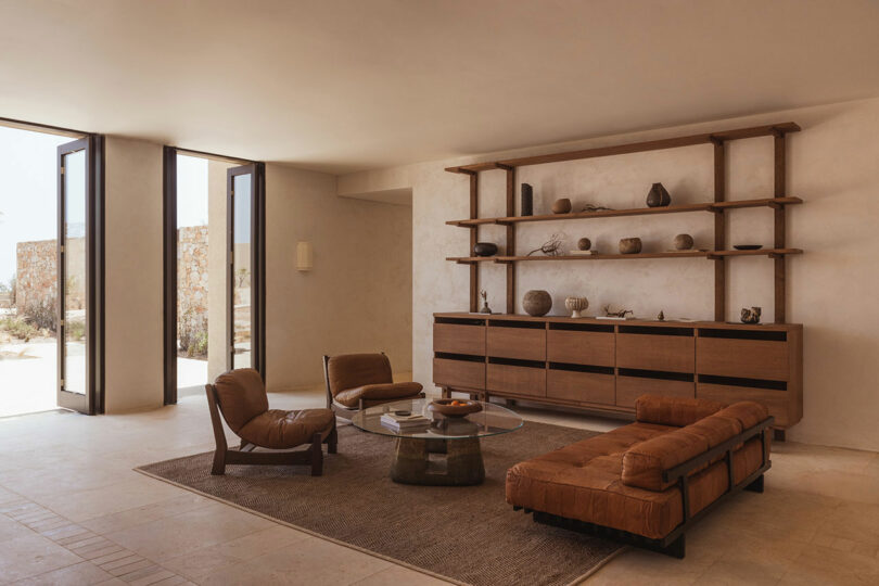 A minimalist living room features brown leather seating, a glass coffee table, and a wooden shelving unit with decorative items on a neutral wall. Large windows provide ample natural light.