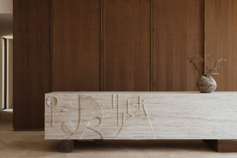 A minimalist lobby area featuring a large stone reception desk with carved abstract designs and a vase with dry branches, against a backdrop of tall, dark wooden panels.