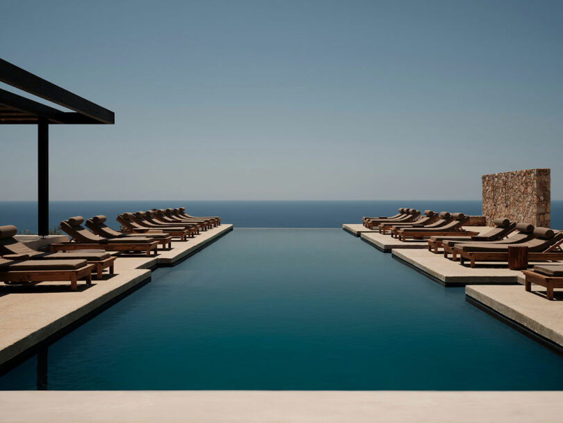 Infinity pool with wooden lounge chairs arranged on each side, overlooking a calm ocean under a clear blue sky.