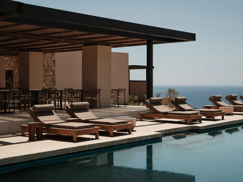 Outdoor poolside area with wooden loungers and a shaded seating area, showcasing a view of the ocean in the background under a clear sky.