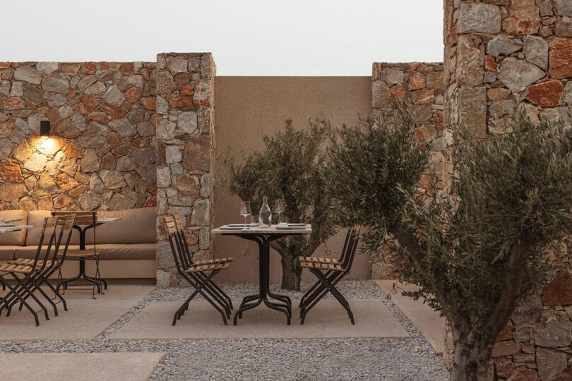 Outdoor patio with stone walls features a small round table with two chairs, a bench with cushions, a wall light, and several potted plants. The setting appears serene and inviting.