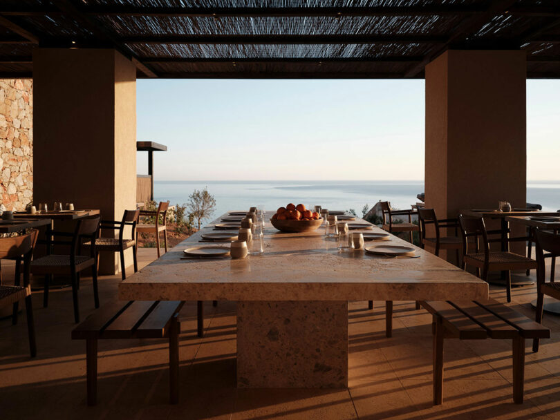 Outdoor dining area with a large stone table set with dishes, benches on either side, and several chairs; overlooks a serene ocean view at sunset.