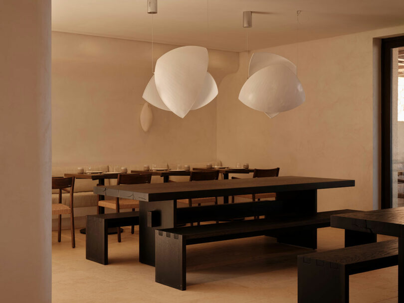 A minimalist dining area with wooden tables and chairs, adorned with unique white pendant lights. The setting has a neutral color palette and a serene ambiance.