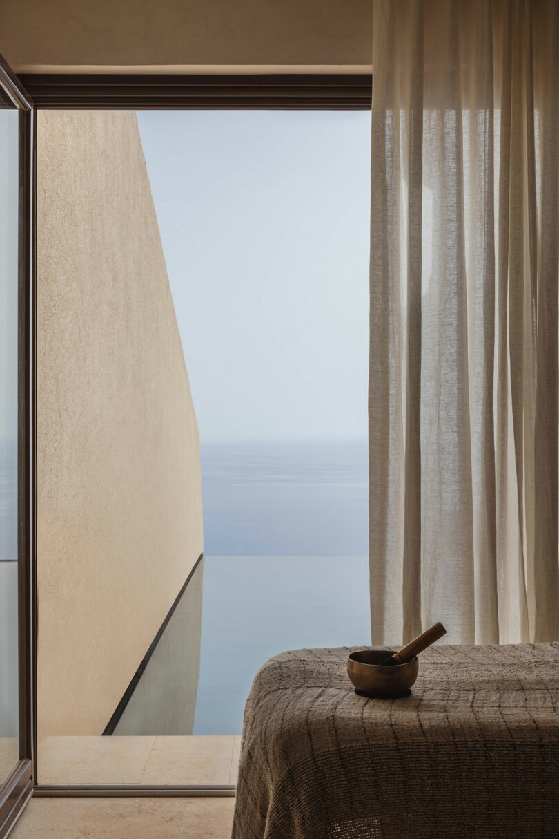 A serene room with sheer curtains partially open, revealing a minimalist infinity pool and a seamless ocean view through a large window. A wooden bowl and pestle sit on a textured table in the foreground.