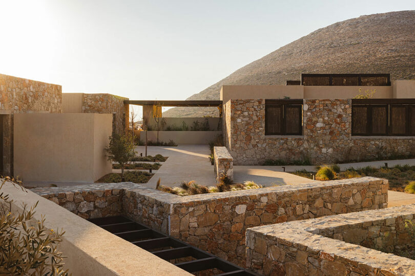 A modern structure built with stone and concrete, featuring several interconnected buildings and walls, is set against a backdrop of rolling hills under a clear sky.