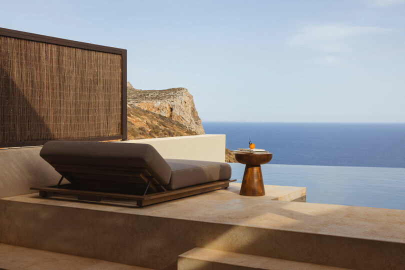 A modern patio with a cushioned lounge chair and a small table overlooking a calm sea and distant cliffs.