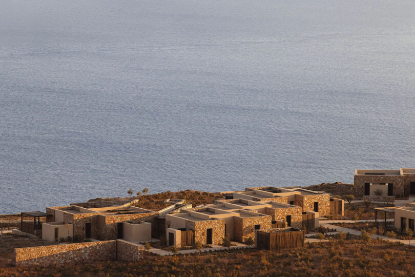 Stone buildings with flat roofs arranged in a pattern near a calm sea, surrounded by dry vegetation.