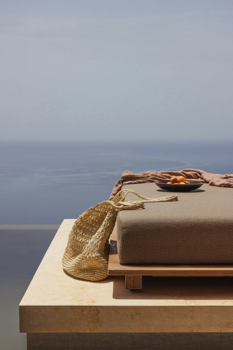 A woven bag and a plate with fruit are placed on a lounge chair overlooking a calm sea under a clear sky.