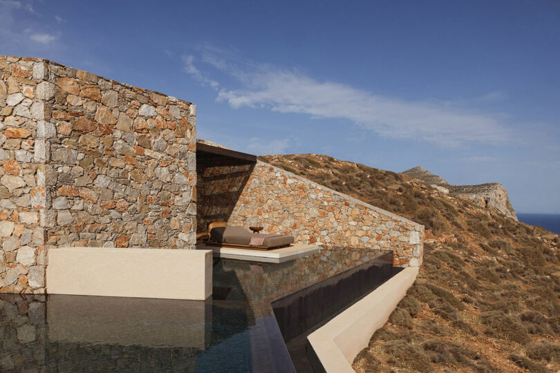 Stone-walled building with flat roof and minimalistic design overlooking rocky terrain and sea in the distance, with an infinity pool in the foreground under a clear sky.