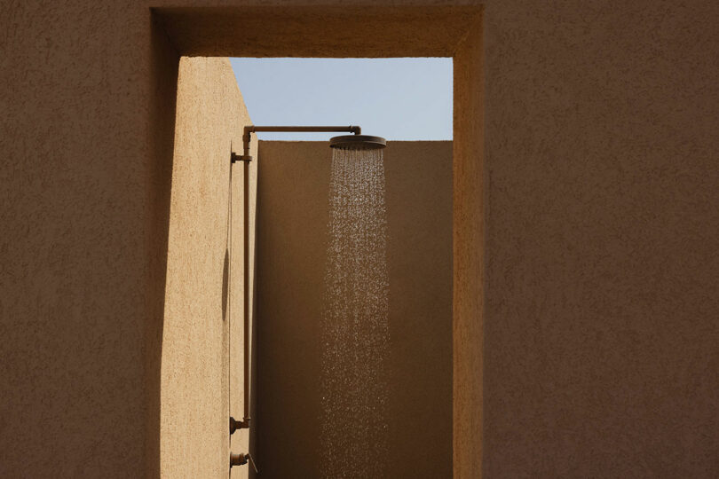 An outdoor shower with water falling from a showerhead mounted on a beige stucco wall, framed by a rectangular doorway, under clear sky.