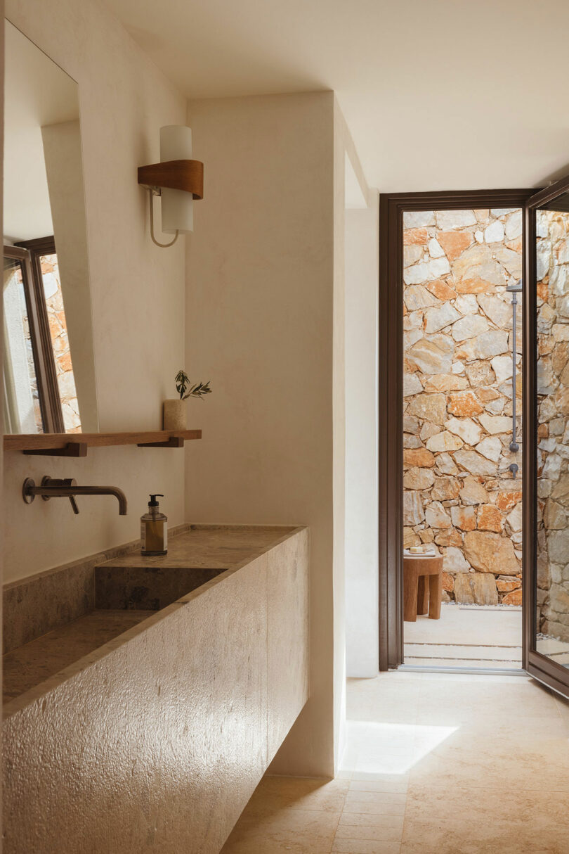 A minimalist bathroom features a stone sink with a wall-mounted faucet, a mirror, and a small shelf. Natural light enters through a door opening to an outdoor stone wall.