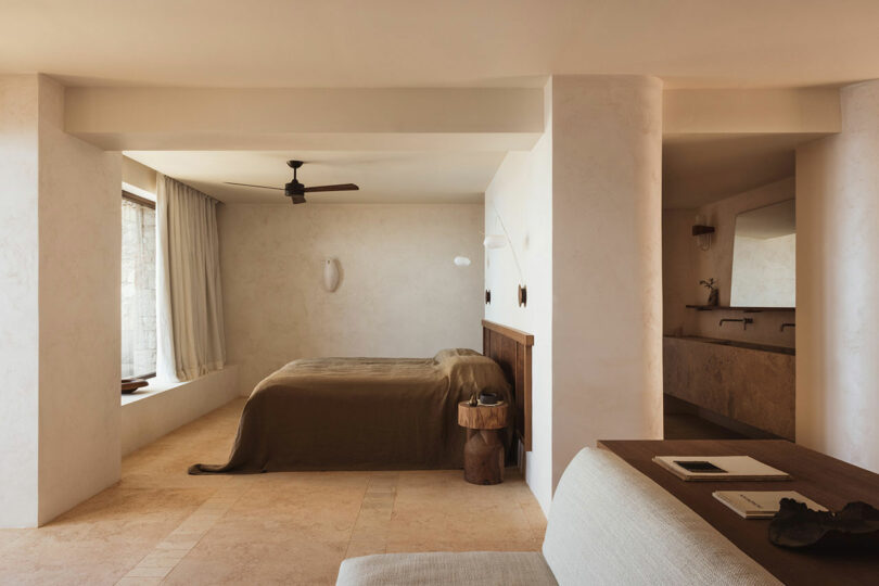 A minimalist bedroom with a beige color scheme, featuring a bed with brown linens, a ceiling fan, and a wall-mounted light. A bathroom with a double sink is visible in the background.