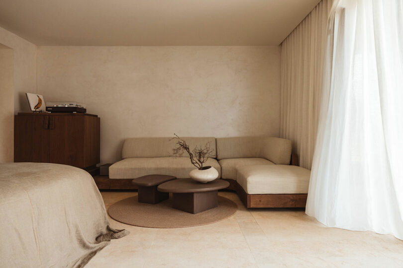 A modern living room with a beige L-shaped sofa, two round coffee tables, a minimalist decor vase, and a record player on a cabinet. Sheer curtains cover a large window, and a bed is partially visible.