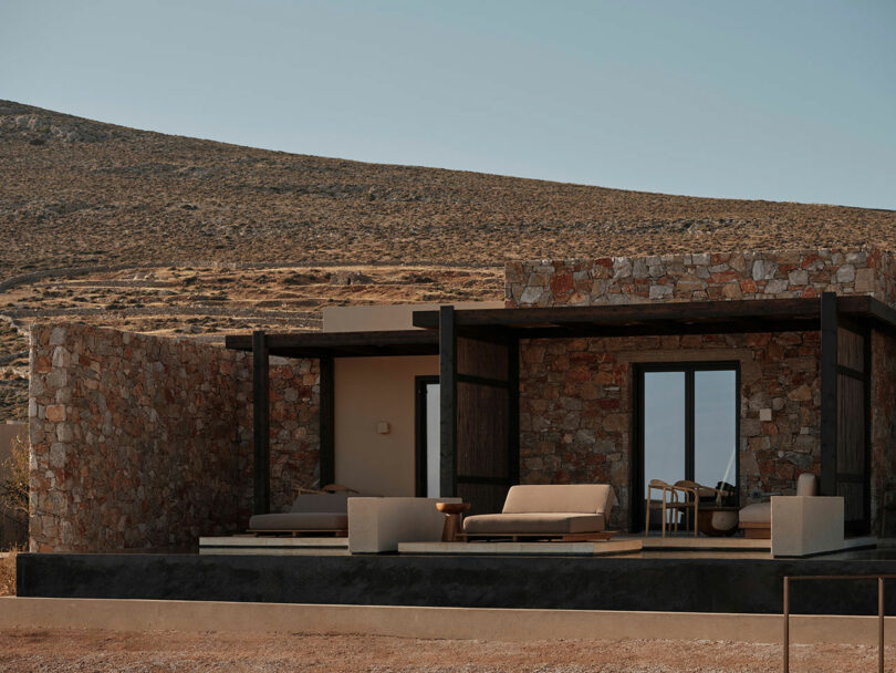 Gundari stone house with a flat roof, large windows, and a covered outdoor seating area in a dry, rocky landscape.