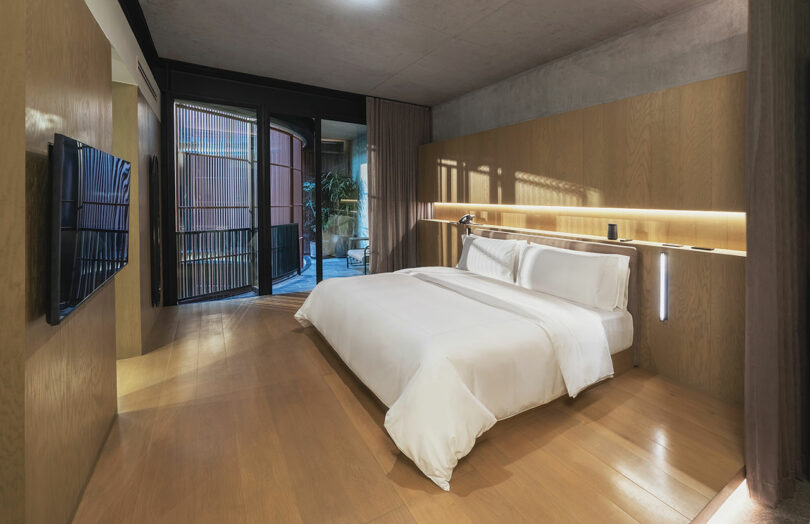 A modern bedroom at Hotel Volga features a large bed, wooden flooring, a wall-mounted TV, floor-to-ceiling windows, and sliding glass doors leading to an outdoor area.