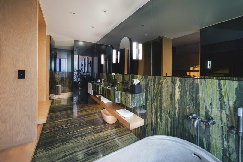 Enjoy a luxurious stay at Hotel Volga, featuring a modern bathroom with green marble walls, a double sink vanity with mirrors, recessed lighting, wooden elements, and a freestanding bathtub.