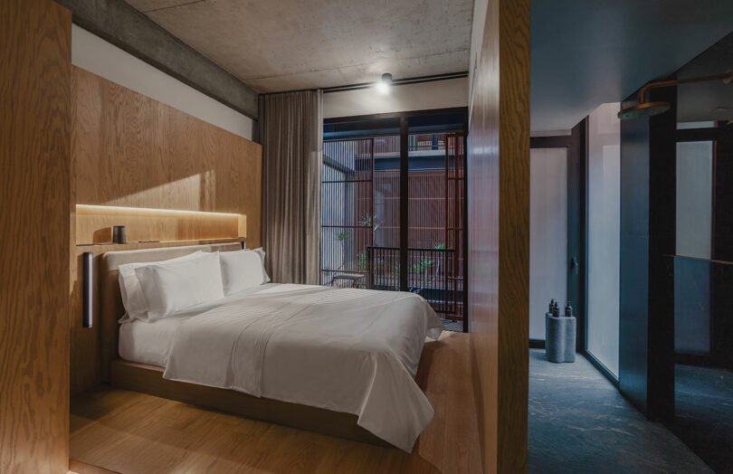 A modern bedroom at Hotel Volga features a wooden accent wall, a neatly made bed with white linens, floor-to-ceiling curtains, and a view of the outdoor area through a glass door.