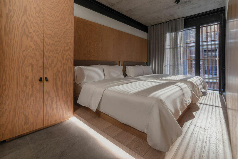 A bedroom at Hotel Volga with two neatly made beds, wooden walls, a wooden wardrobe, and large windows with sheer curtains allowing natural light.