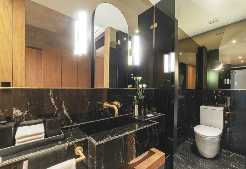 A modern bathroom at Hotel Volga featuring a black marble sink, brass fixtures, a mirror with vertical lights, a toilet, and wooden accents.