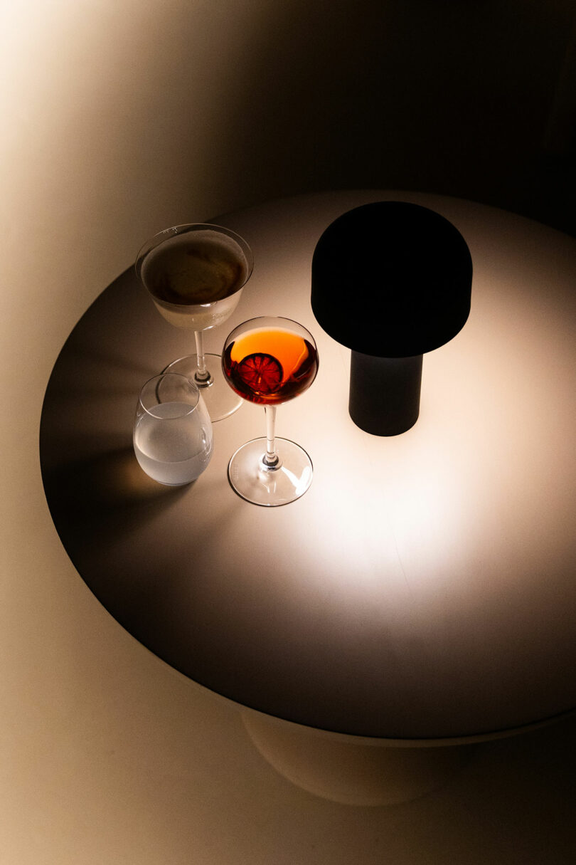 A round table at Hotel Volga holds three drinks: a glass of water, a martini, and a cocktail with an orange slice. A dark lamp stands next to the drinks, illuminating the scene.