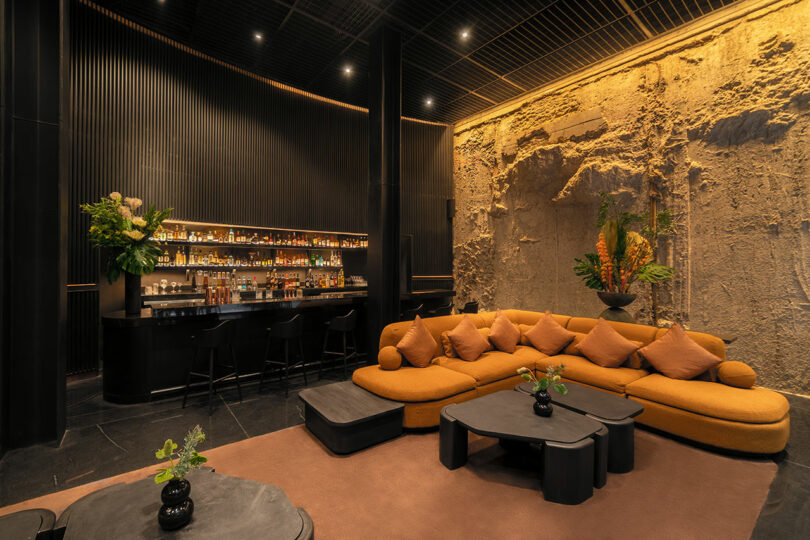 A stylish lounge at Hotel Volga features a curved bar, black stools, and a mustard-colored sectional sofa. The walls are a blend of textured stone and sleek black surfaces, while tables are adorned with plants.