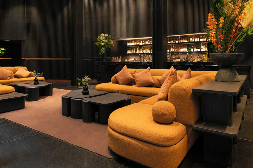 The stylish lounge area at Hotel Volga features mustard-colored sofas with various cushions, black tables, and a well-stocked backlit bar in the background. The decor includes green and orange floral arrangements.