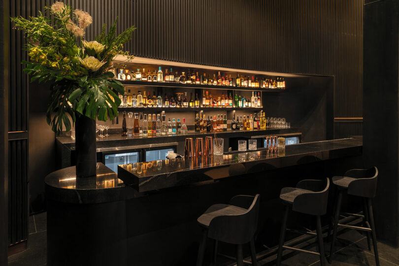 A dimly lit modern bar at Hotel Volga features a black countertop, three stools, various bottles displayed on shelves, and a floral arrangement on the counter.