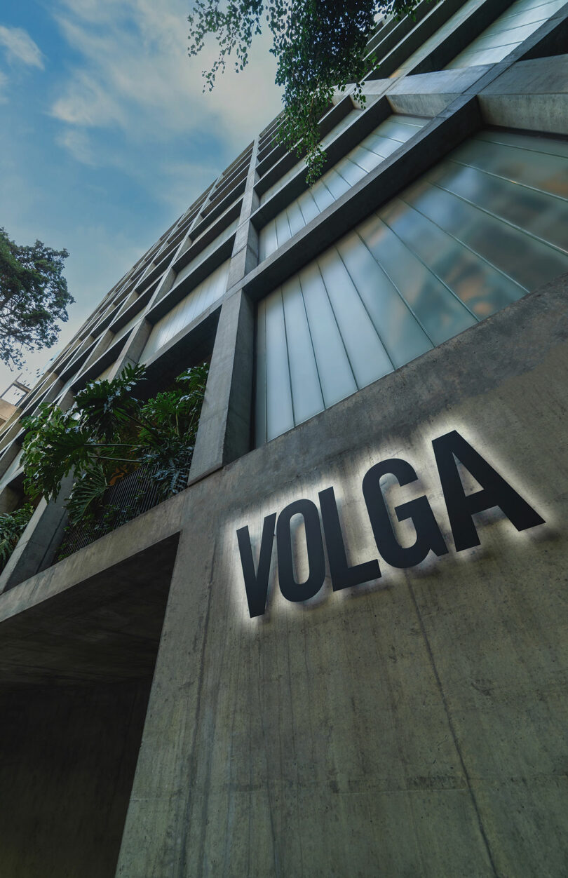 A modern high-rise building seen from a low angle with the name "Volga" prominently displayed on its concrete facade. The structure features large windows and visible greenery.