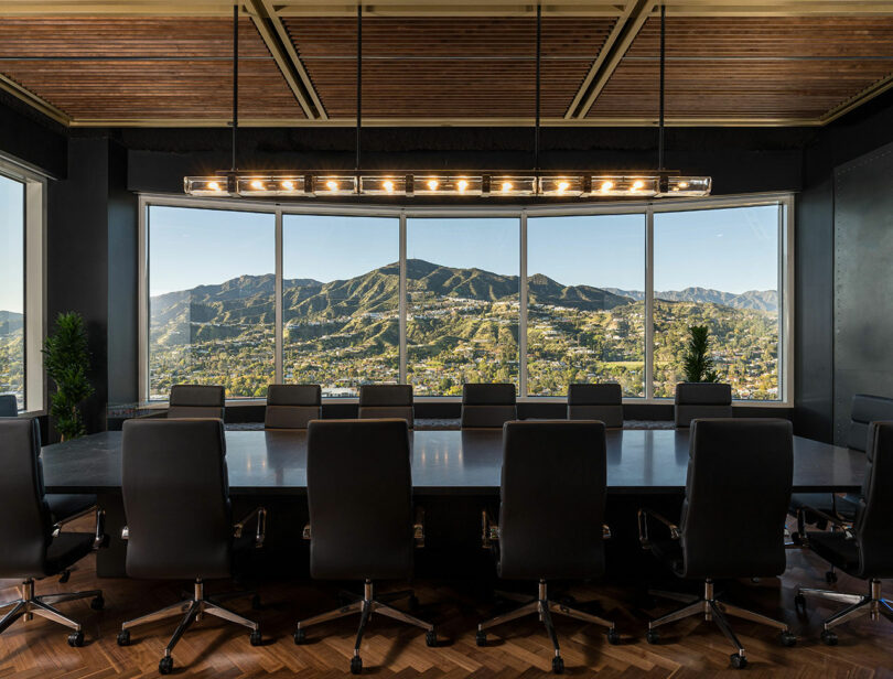 A modern conference room with a large table and black chairs, featuring a panoramic window with a view of a mountain range.