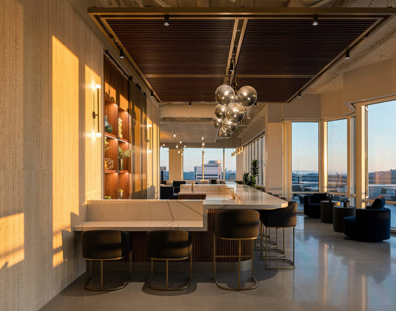 Modern lounge area with marble countertops, bar stools, and a large contemporary chandelier. Sunlight streams in through the large windows, highlighting the sleek interior design and city view.