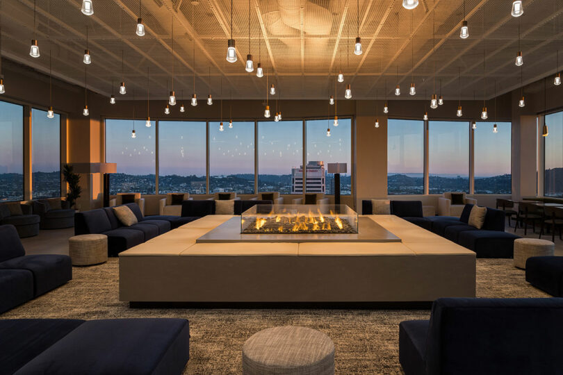 A modern indoor lounge with a central gas fireplace, surrounded by seating, large windows offer a panoramic view of a cityscape during twilight.