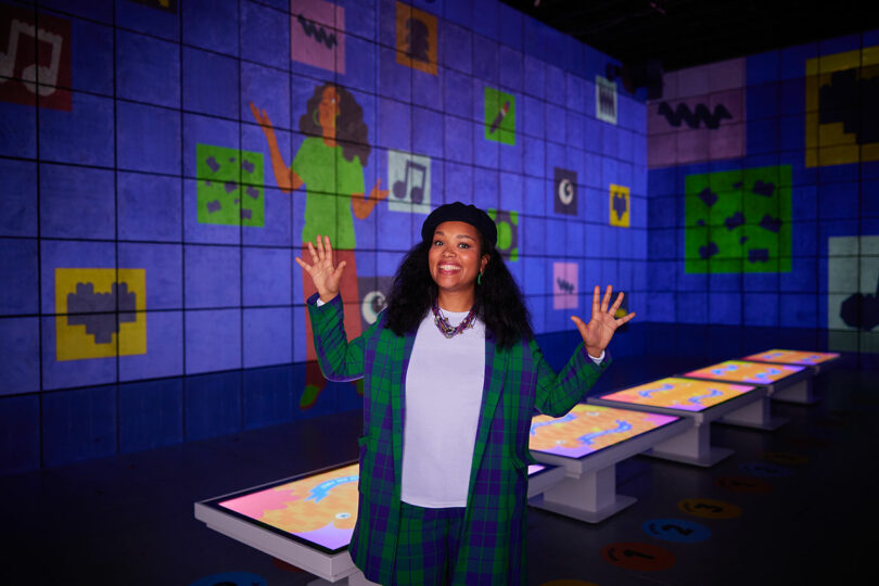 A person wearing a green and blue checkered outfit and black hat stands in a colorful, futuristic room with wall projections and touch-screen tables. They are smiling and gesturing with raised hands.