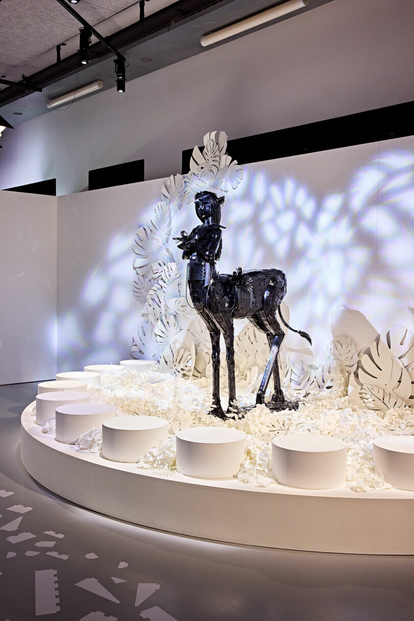 A black centaur statue stands in an art exhibit surrounded by white sculptural elements and lighting effects.