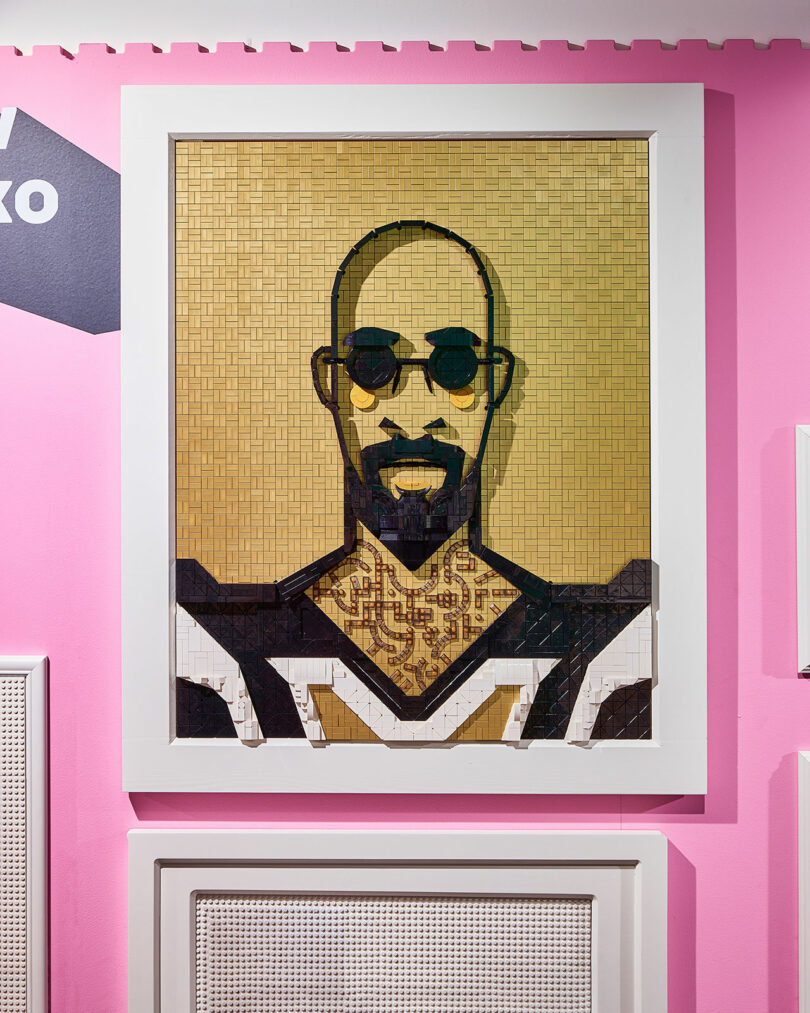 A framed mosaic portrait of a bald man with a beard and sunglasses against a pink wall.