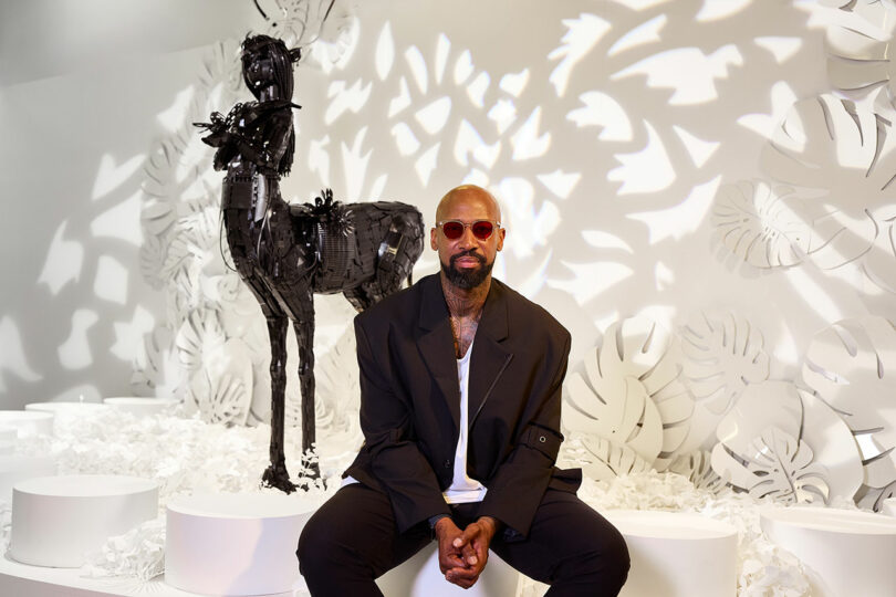 A man in a black jacket and red sunglasses sits in front of a black centaur sculpture with a white floral abstract background.