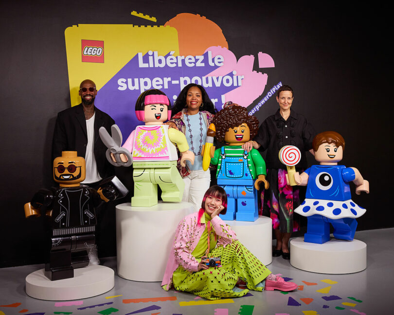 Four people stand behind life-sized LEGO figures, with a colorful backdrop that includes the French phrase "Libérez le super-pouvoir." Confetti decorates the floor, and everyone appears cheerful.