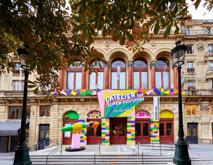 Colorful, vibrant entrance to an art exhibit with playful and whimsical decorations on a historical building facade, framed by leafy tree branches and streetlights.