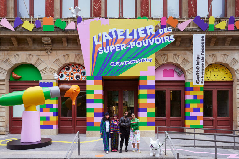 Three people stand in front of a colorful, playful entrance titled "L'ATELIER DES SUPER-POUVOIRS" with a sign reading "#SuperpowerofPlay." A large dog sculpture is also present by the steps.