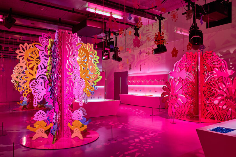 A brightly lit room with colorful, intricate paper cut-out displays in pink, red, and orange hues, alongside shelves illuminated with pink lighting. The space has a whimsical, artistic ambiance.