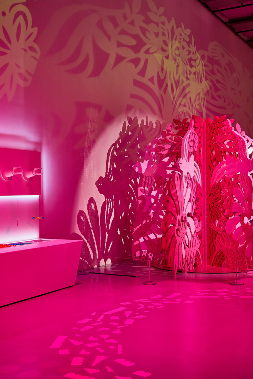 A room illuminated with pink light features an intricate floral-patterned screen and a modern white desk with colorful objects. Shadows of the floral pattern are cast on the walls and floor.