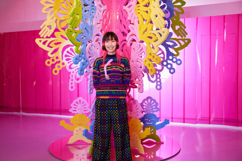 A person stands smiling in front of a colorful, intricate art installation with pink background and patterned floor. The person wears a multicolored striped sweater and matching wide-legged pants.