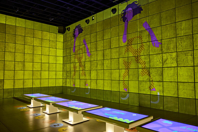 A room with tiled walls displays cartoon images of a person in motion. Interactive touchscreens are mounted on stands in front of the walls. The room is lit with greenish lighting.