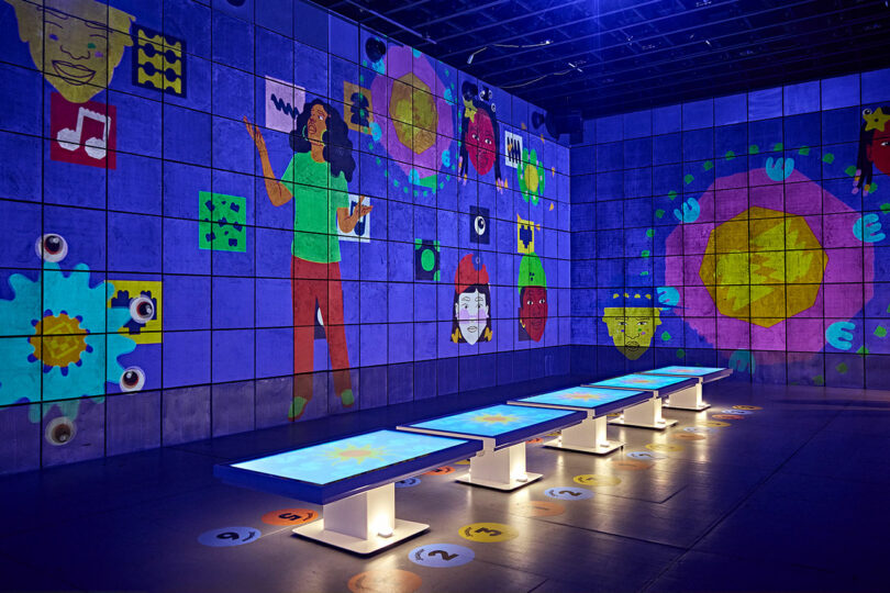 Interactive exhibit with illuminated tables in front of large colorful wall displays featuring various graphics and portraits. The area is dimly lit with a focus on the bright elements.