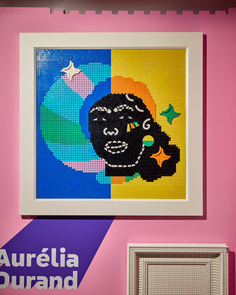 Framed colorful mosaic artwork on a pink wall with sections of blue, green, and yellow, featuring a stylized face. Text "Aurélia Durand" is partially visible in the lower left corner.