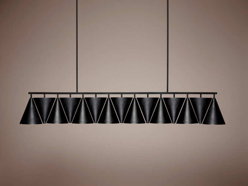 A Legato Linear by Luca Nichetto - modern black pendant light fixture with a row of conical lampshades, suspended from a ceiling by two rods against a neutral background.