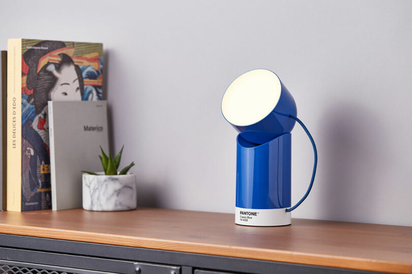 A blue modern lamp labeled "Pantone" on a wooden shelf, next to a small potted plant and several books.