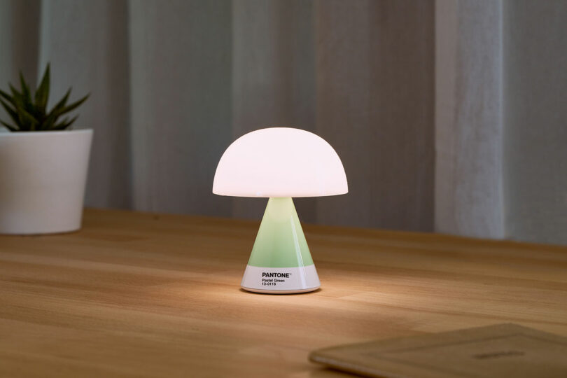 A small, dome-shaped LED table lamp with a green gradient sits on a wooden surface, next to a plant in a white pot and a beige notebook.