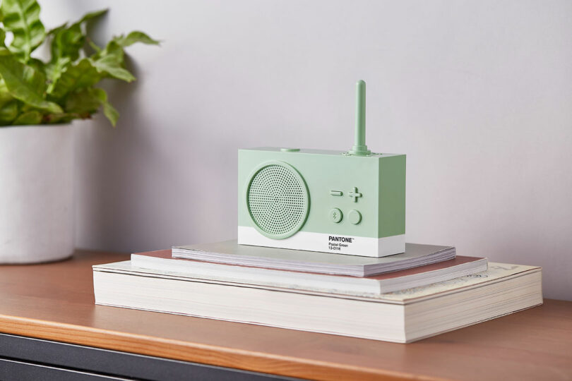 A green and white tabletop radio with an antenna, featuring Pantone branding, is placed on top of stacked books beside a potted plant on a wooden surface.